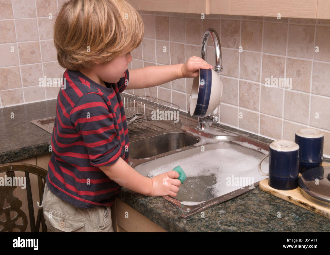 young child washing the dishes at the kitchen sink standing on a stool with the hot tap running Stock Photo