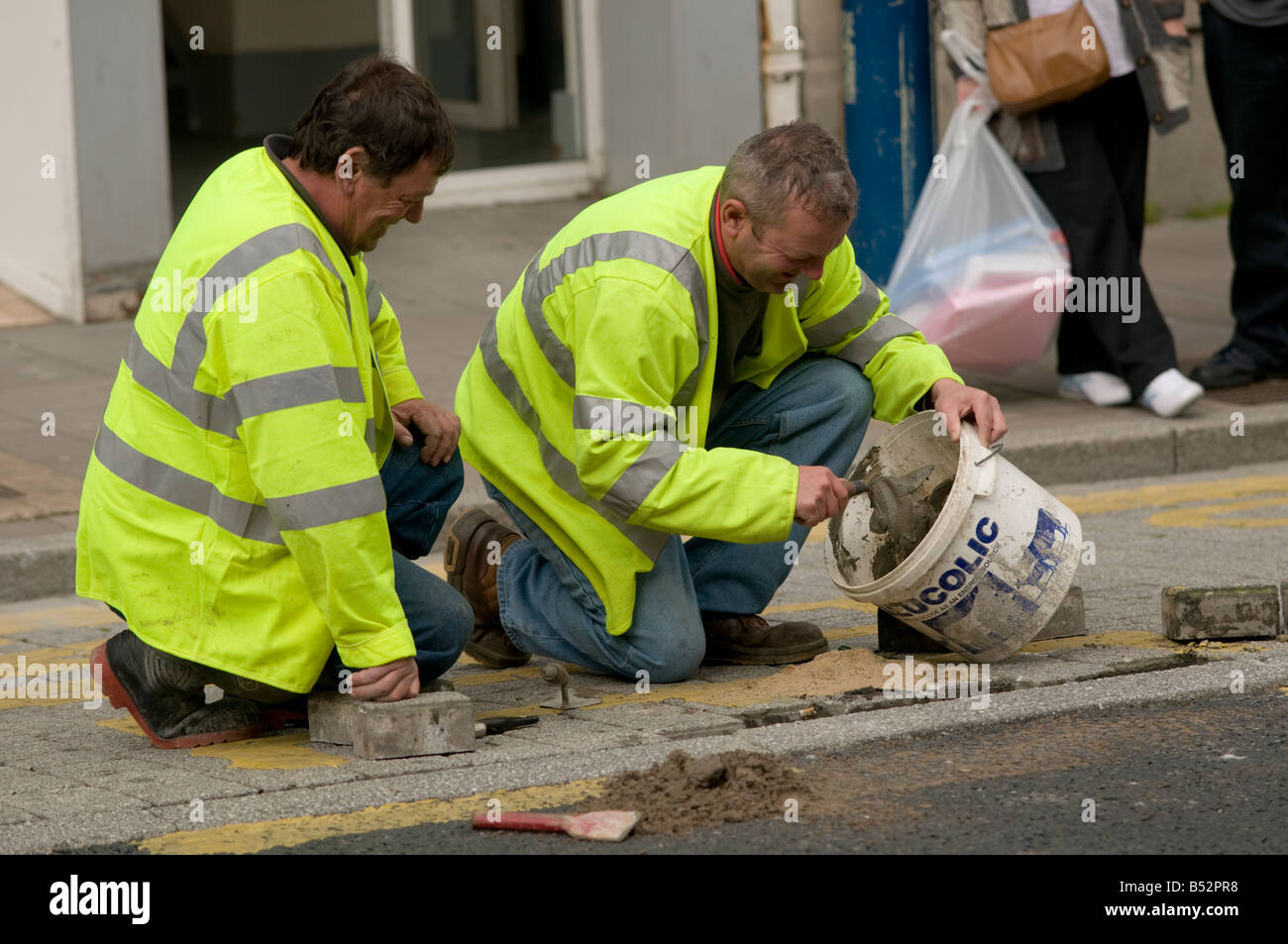 Two local authority workers repairing pavement wearing bright yellow high visibility jackets kneeling down, UK Stock Photo