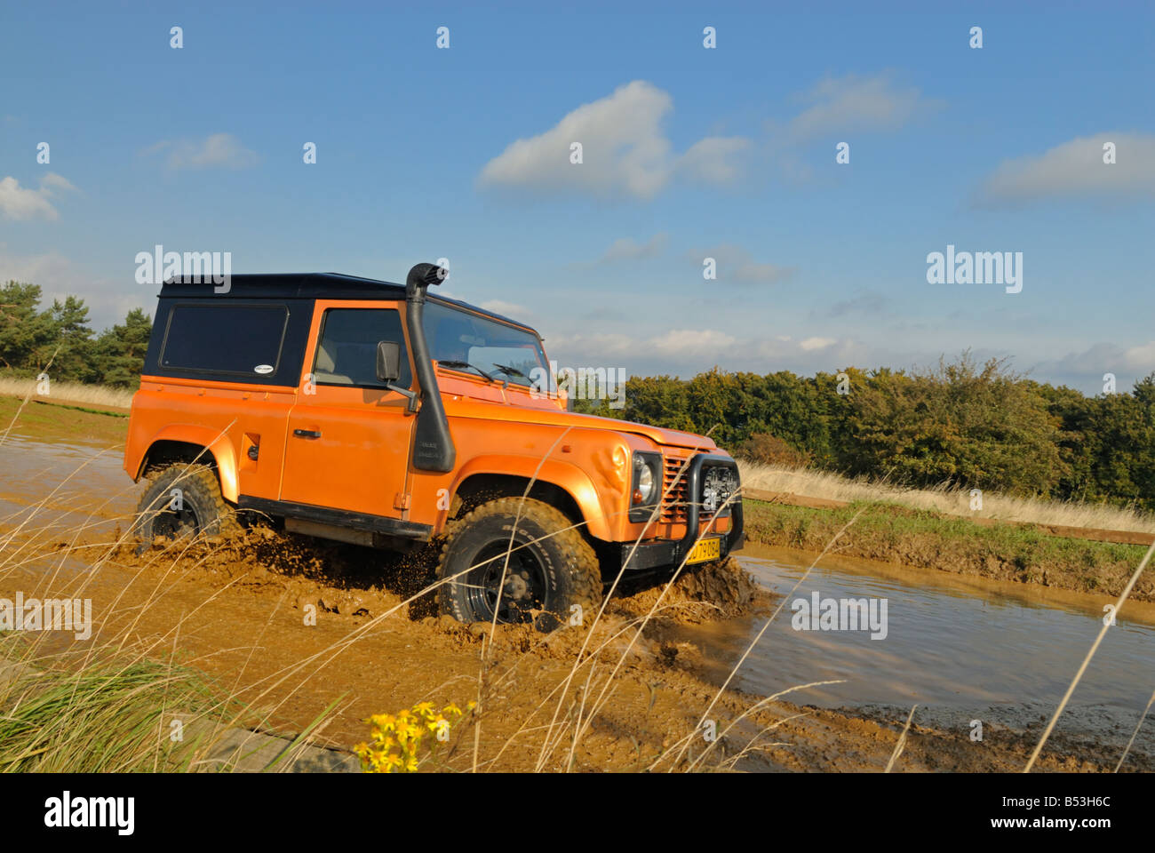 Orange coloured Land Rover Defender 90, fitted with a high raised air intake, crossing a flooded forest track. Stock Photo