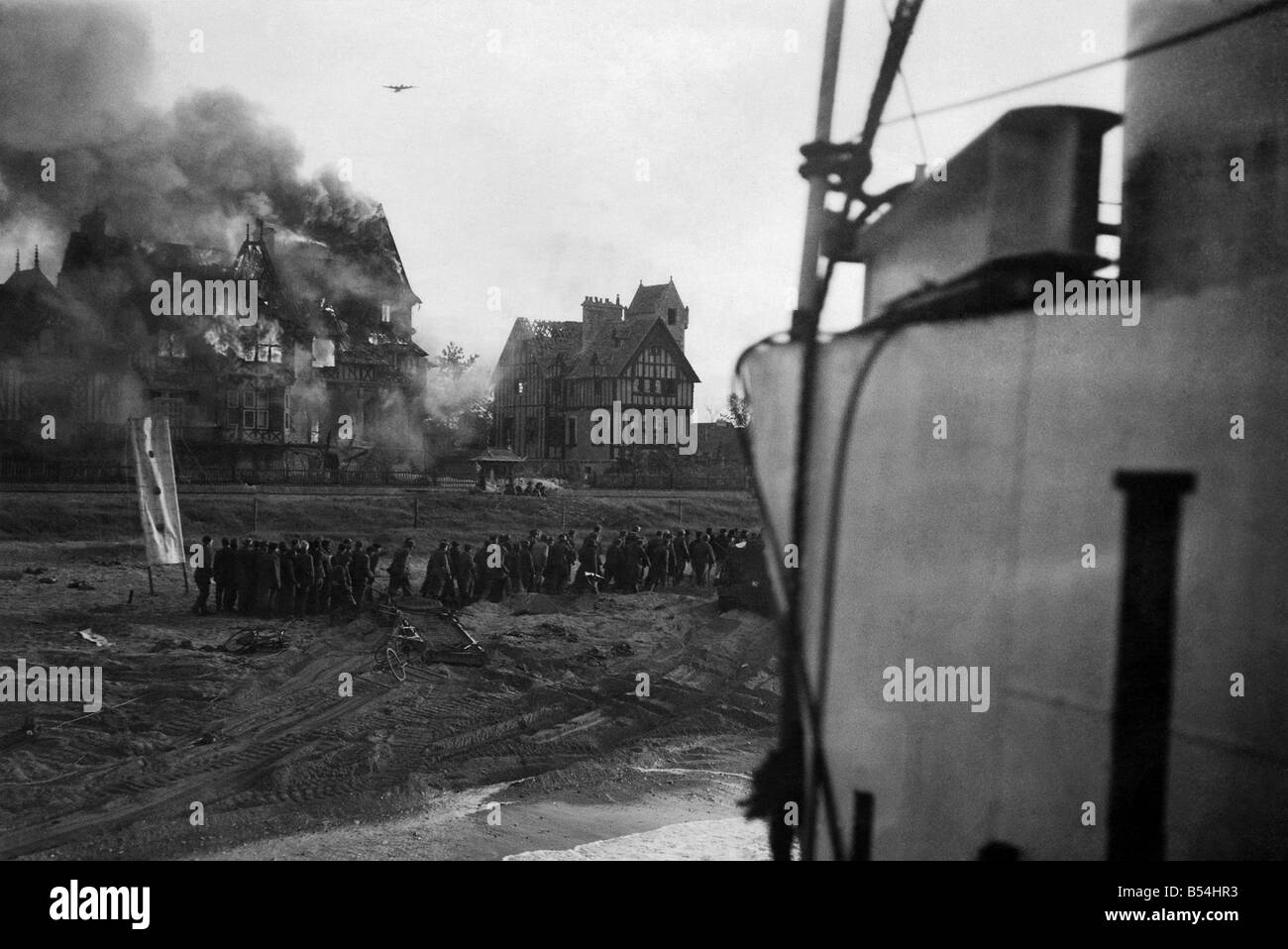 British troops under fire on Juno beach at Normandy shortly after the D-Day landings. ;Soon after zero hour German aircraft seen in sky over beach. Has just bombed house seen in flames in the foreground a column of German prisoners just captured. June 1944 ;P011596 Stock Photo