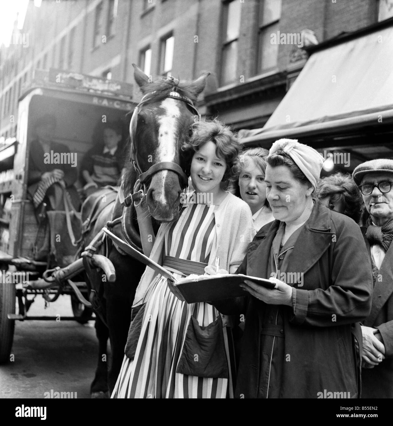 Children petition to save milkmans house: Jennifer here collects a signature in a street market at Bedford Hill, Balham. Signing the book is Mrs. Robert Kelly who has given him a carrot every morning for years. Jennifer gives Eva a carrot on the right is Diane Cathercole who has helped gather signatures. ;June 1960 ;M4407 Stock Photo