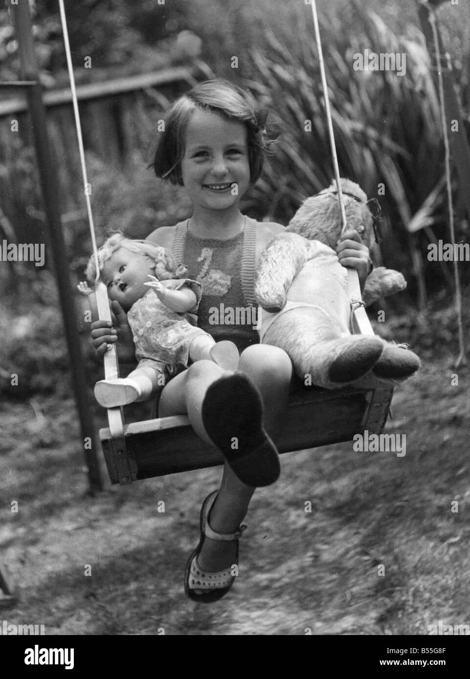 Young girl sitting on her swing in the back garden with her favourite 'toys,' a doll and teddy bear on either side Circa 1945 Stock Photo