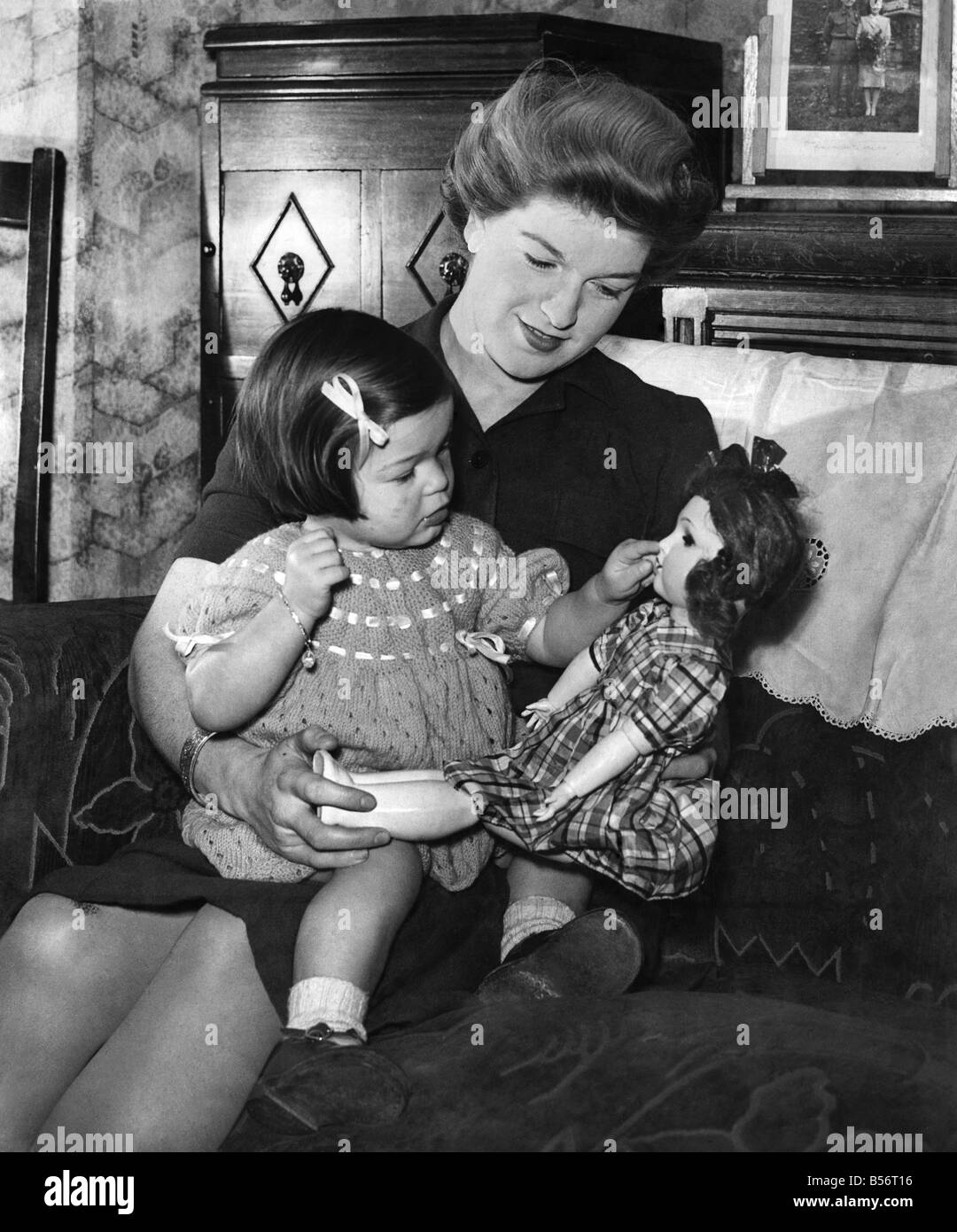 Mother and child at home, the girl playing with her doll as she sits on ...
