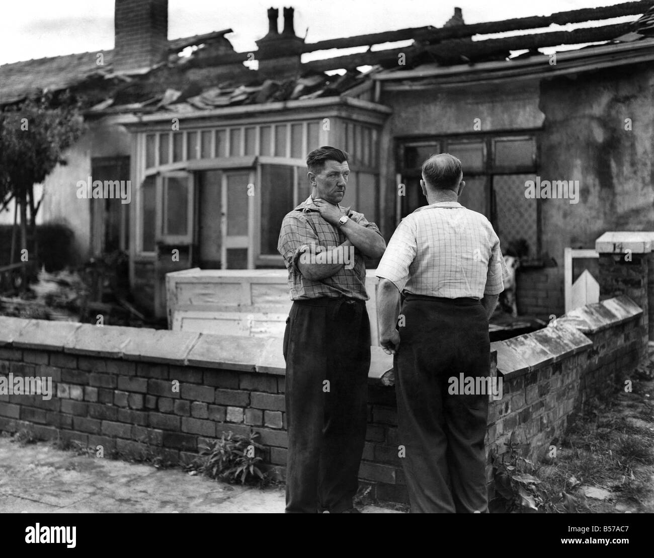 Fireman Jack Skeats, 48, of the Bury Fire Brigade chats to a friend in front of his burnt out bungalow in Hillcrest Ave., Heywood, Lancs. The house caught fire when he was burning off paint with a blow lamp. July 1960 P007261 Stock Photo