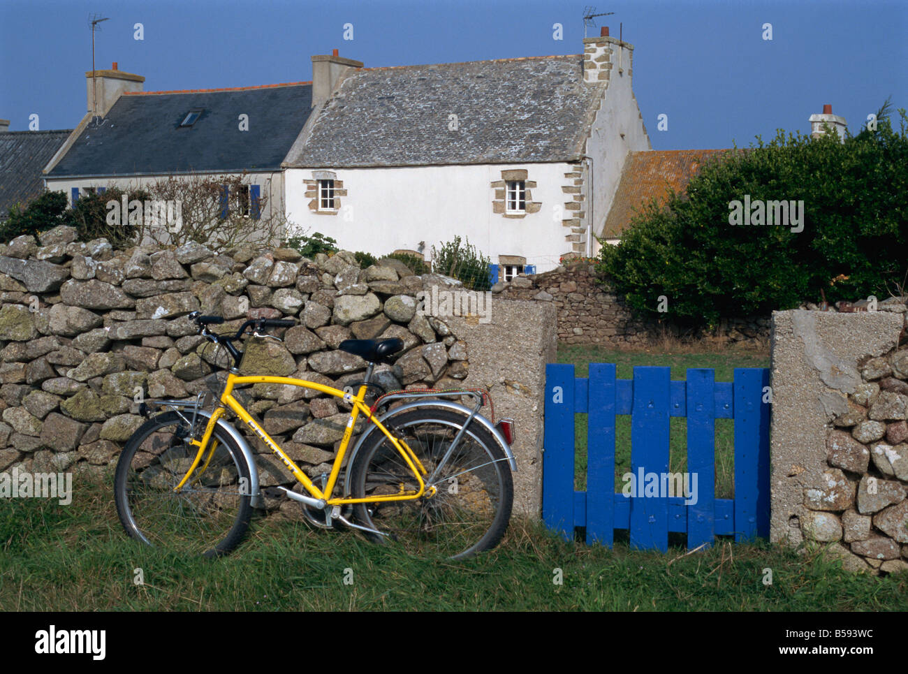 Yellow bicycle beside a blue gate with ancient house behind near Creach lighthouse on Ouessant island Brittany France G Stock Photo