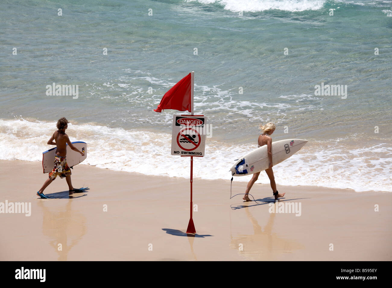 People carrying surf boards past Danger No Swimming sign with red flag on North Stradbroke Island Queensland QLD Australia Stock Photo