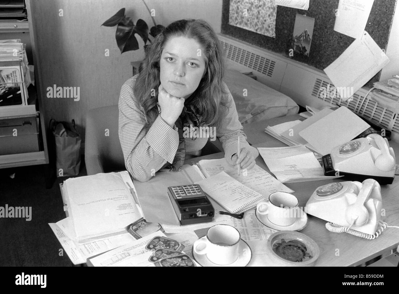 A desk covered with paper, empty coffee mugs and a calculator symbols of the busy, modern executive, and used by attractive Verity Vickers who has moved from being a secretary to an executive with Austin-Toppan Limited. ;February 1975 ;75-01070-001 Stock Photo