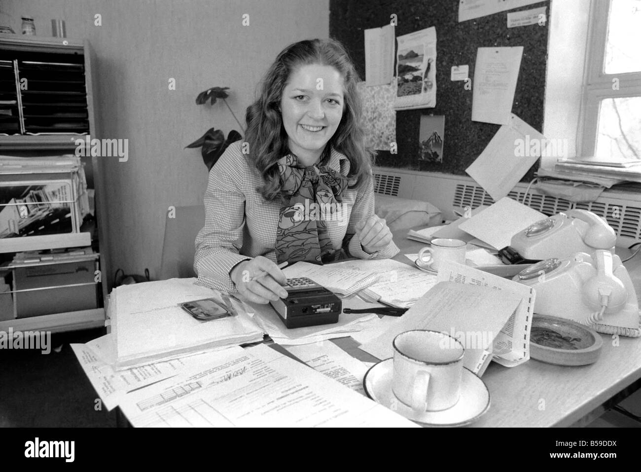 A desk covered with paper, empty coffee mugs and a calculator symbols of the busy, modern executive, and used by attractive Verity Vickers who has moved from being a secretary to an executive with Austin-Toppan Limited. ;February 1975 ;75-01070 Stock Photo