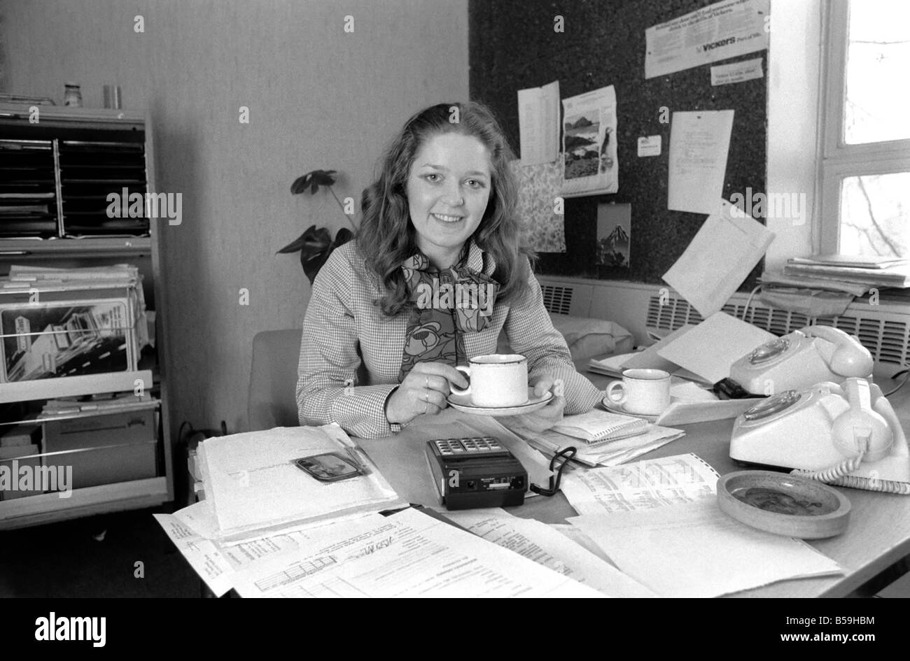 A desk covered with paper, empty coffee mugs and a calculator symbols of the busy, modern executive, and used by attractive Verity Vickers who has moved from being a secretary to an executive with Austin-Toppan Limited. ;February 1975 ;75-01070-002 Stock Photo