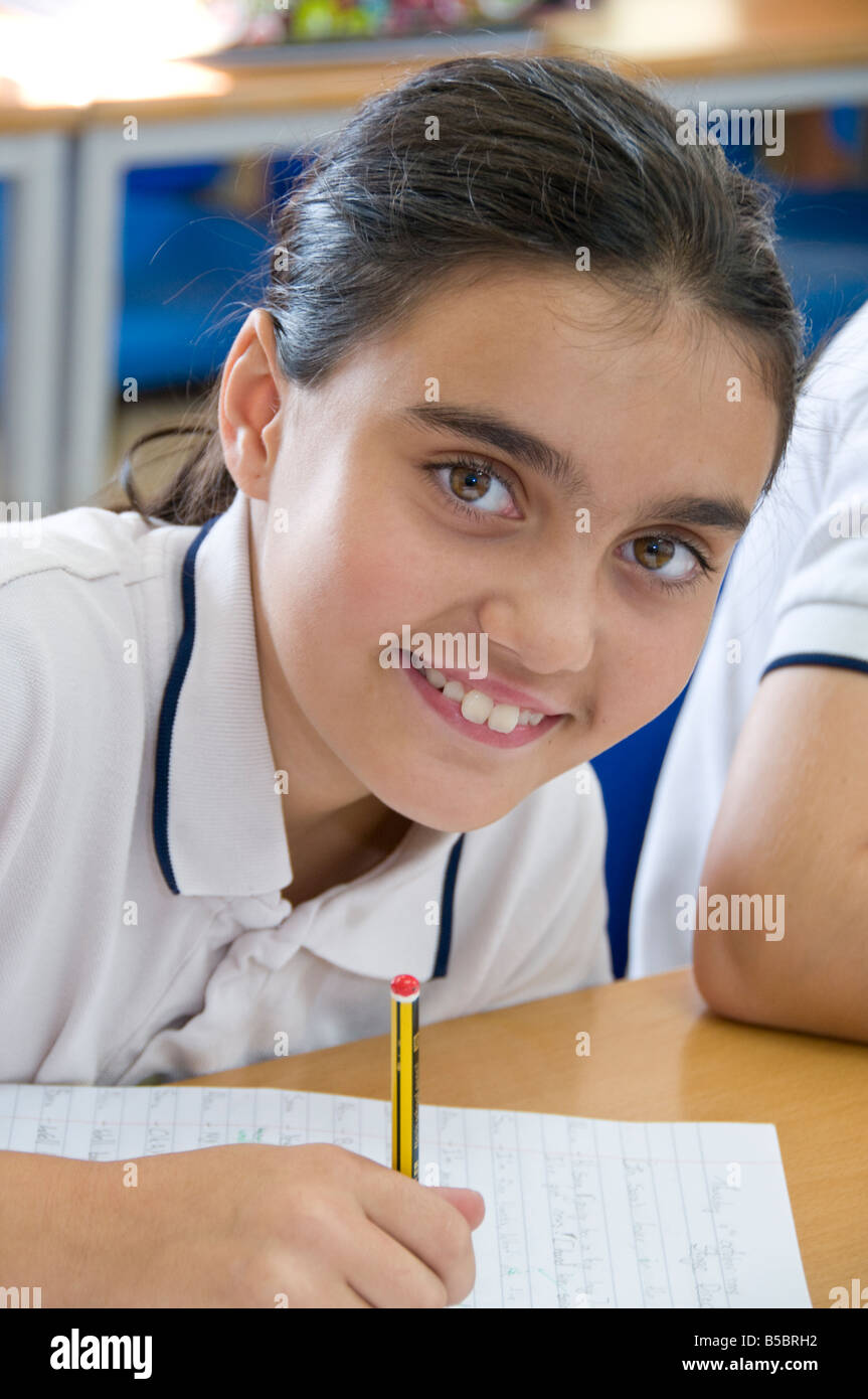 Smiling junior schoolgirl looks up from writing at her desk in school ...