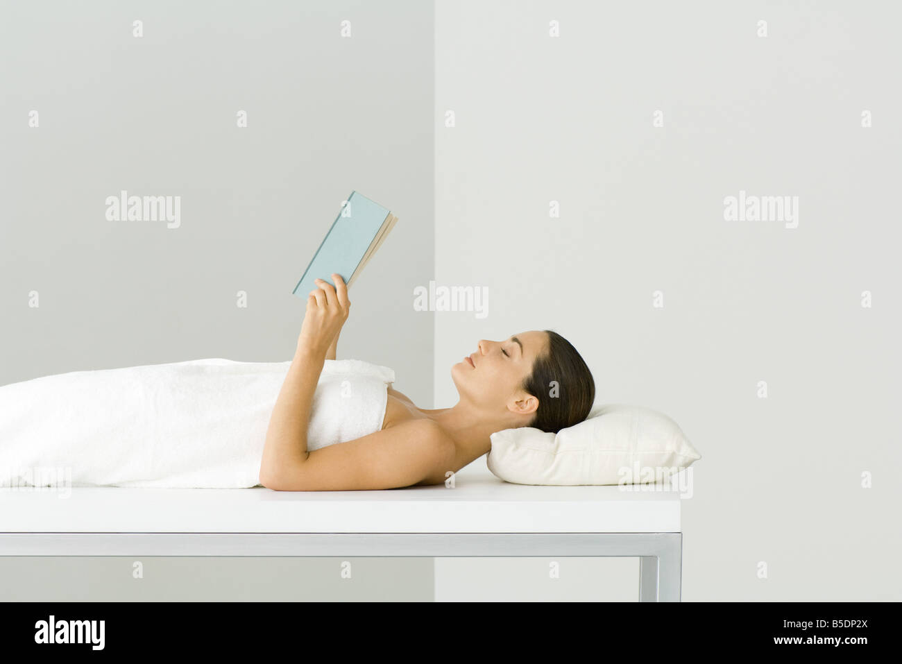 Woman wrapped in towel, lying on massage table, holding book, eyes closed Stock Photo