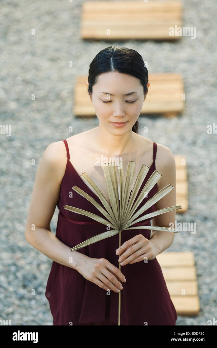 Woman standing in rock garden holding dried palm frond, eyes closed Stock Photo