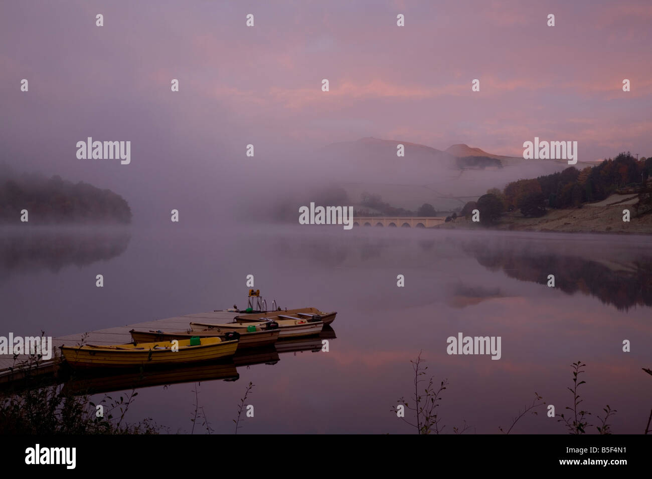 Rowing boats moored on Ladybower Dam with early morning mist rising and red sunlit clouds reflecting in the tranquil water. Stock Photo