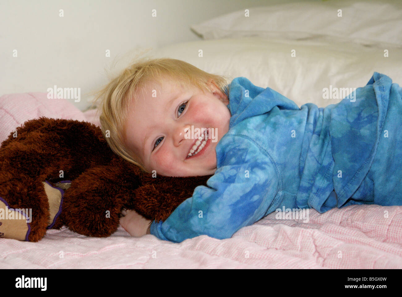 Small blond blue-eyed child dressed in blue  lying on stuffed animal on bed with blurred background Stock Photo