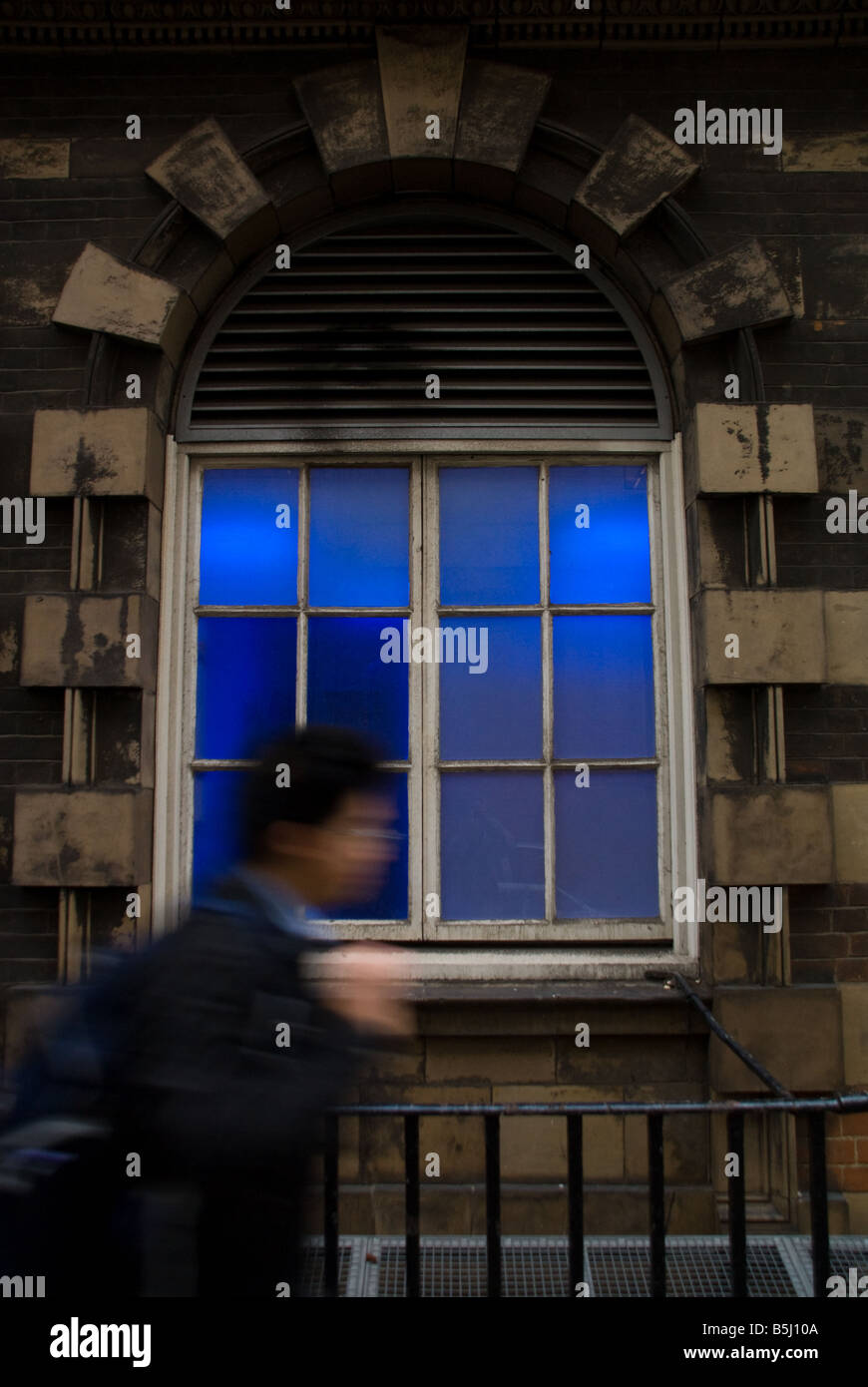 A young asian man walking along a London street past a deep blue arch shaped window Stock Photo