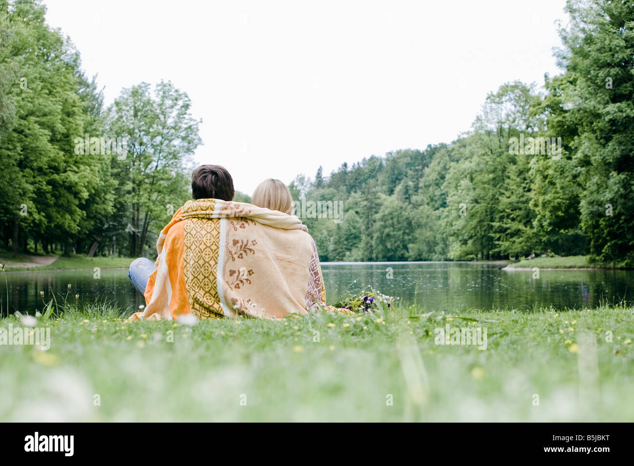 rear view of couple wrapped in blanket sitting at lake in park Stock ...