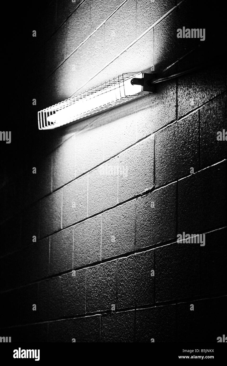 A flourescent light bulb encased by metal housing in a dark and dingy cinderblock wall. Stock Photo