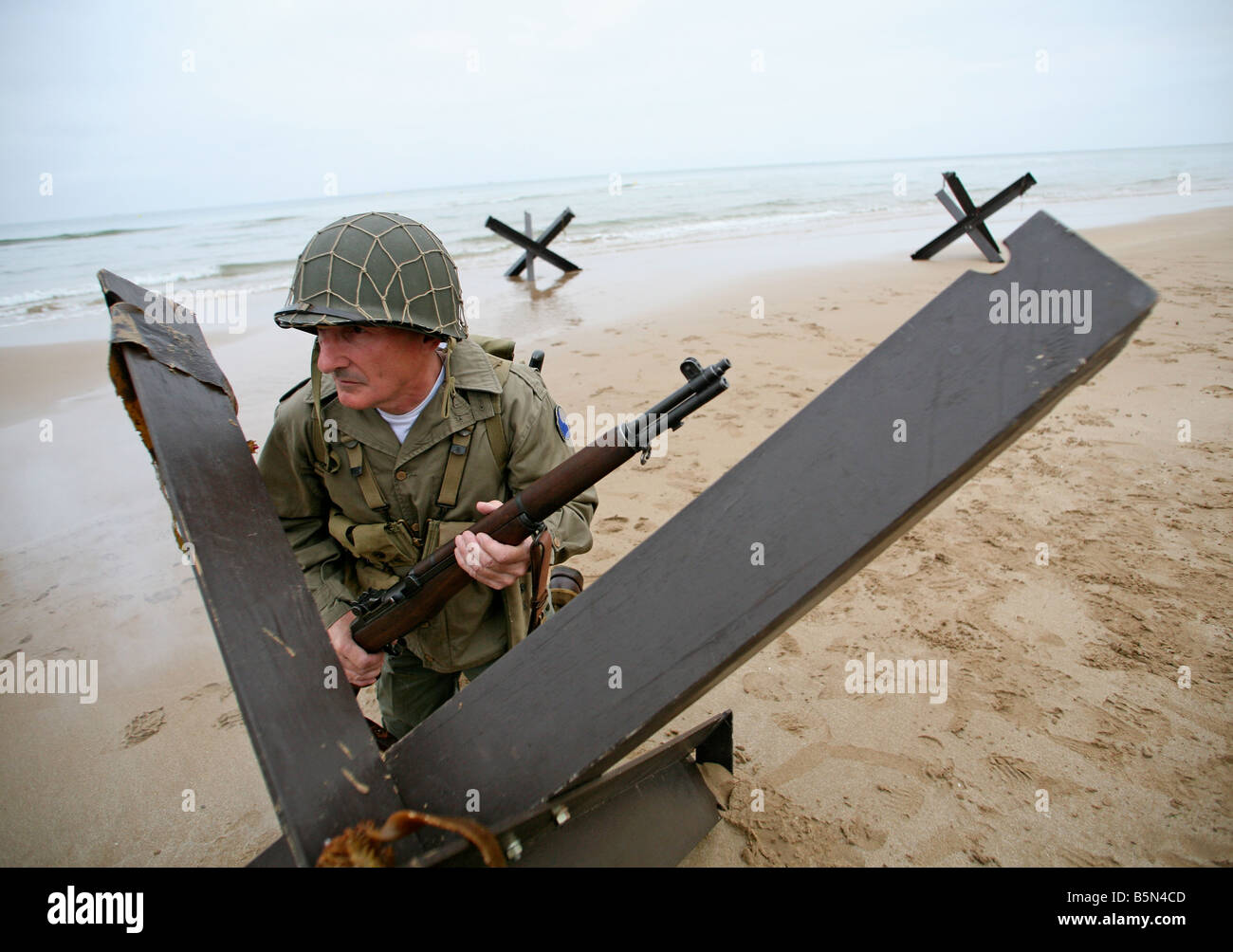 An actor dressed as an D-Day  American soldier on Omaha Beach Normandy France behind a 'Hedgehog' tank trap. Stock Photo