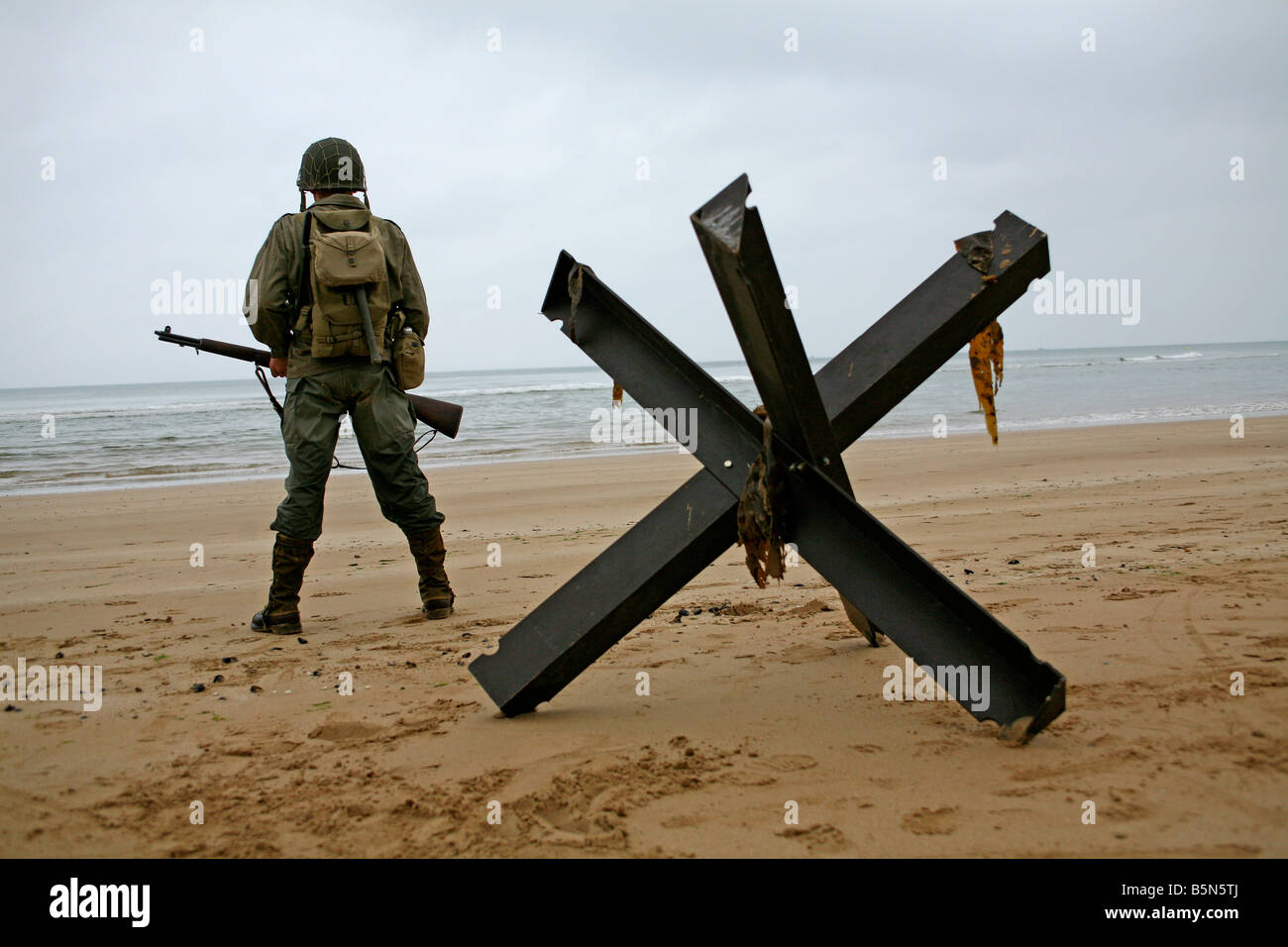 An actor dressed as an D-Day  American soldier on Utah Beach Normandy France standing next to  a 'Hedgehog' tank trap. Stock Photo
