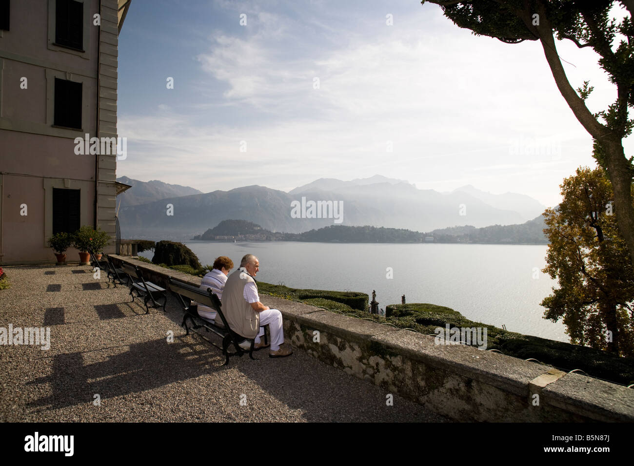 Couple of senior tourists sitting on a bench admiring lake Com view. Villa Carlotta, Tremezzo, Italy Stock Photo