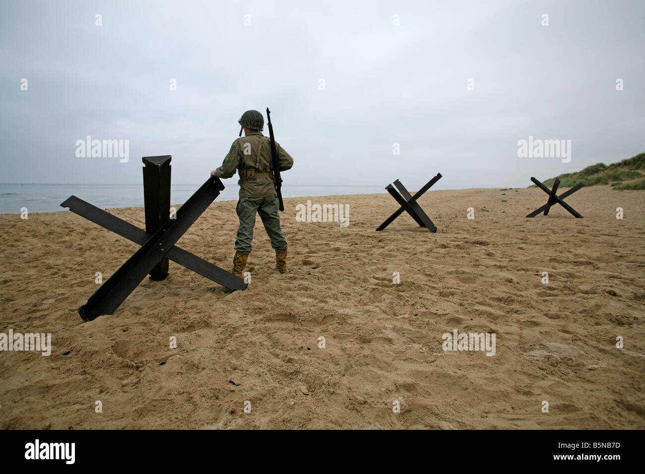 Actor dressed as an D-Day  American soldier on Utah Beach Normandy France standing next to  a 'Hedgehog' tank trap. Stock Photo