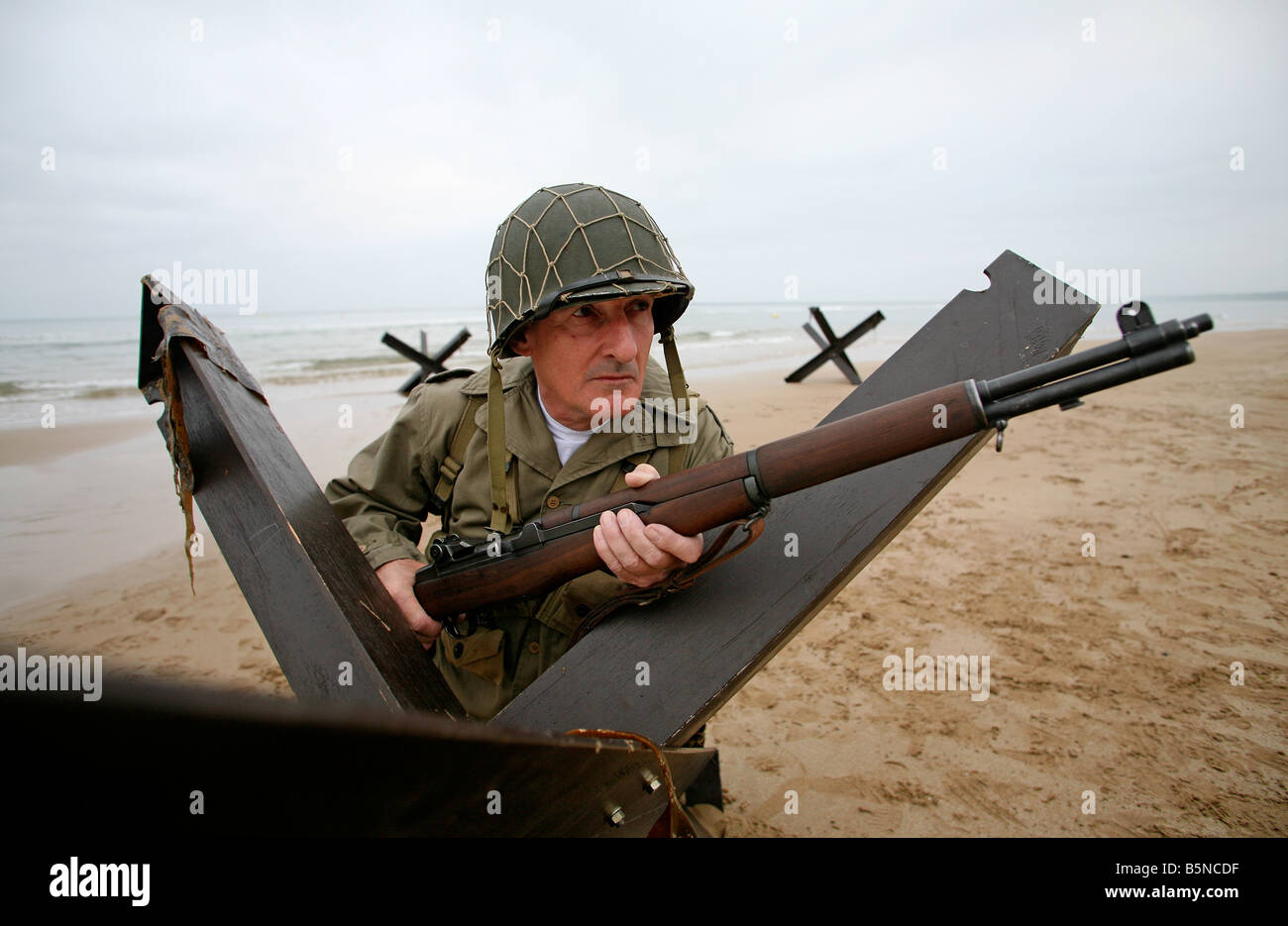 An actor dressed as an D-Day  American soldier on Utah Beach Normandy France behind a 'Hedgehog' tank trap. Stock Photo