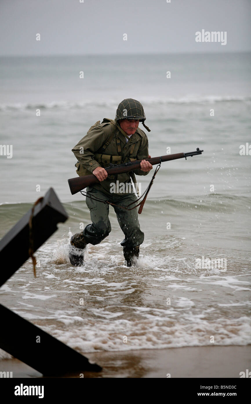An actor dressed as an D-Day  American soldier runs up Omaha Beach Normandy France passed a 'Hedgehog' tank trap. Stock Photo