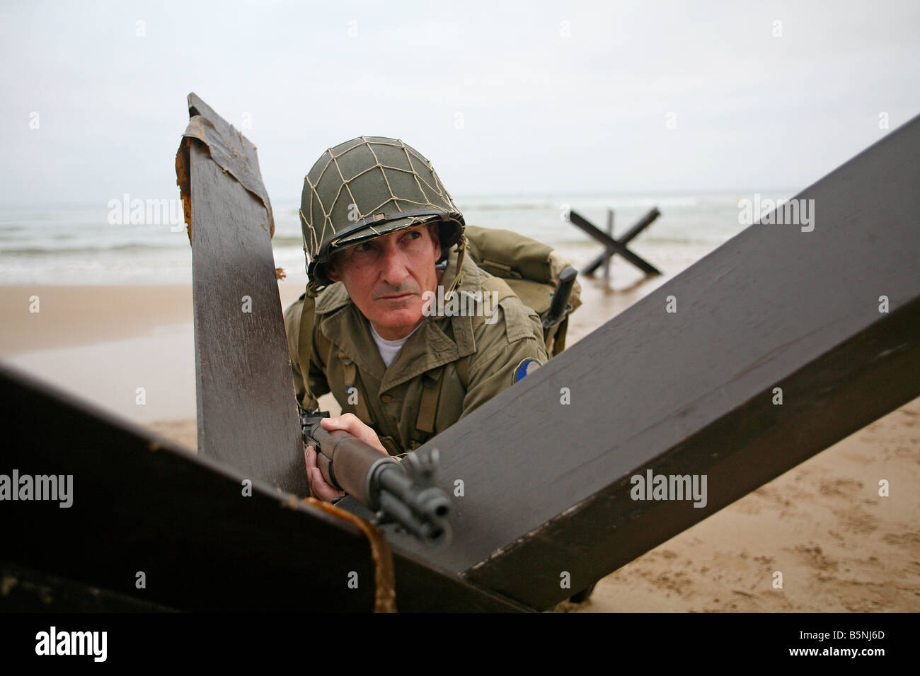 An actor dressed as an D-Day  American soldier on Omaha Beach Normandy France behind a 'Hedgehog' tank trap. Stock Photo