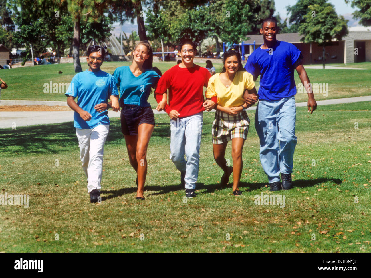 Five ethinic mix teens walk arm in arm across park grass Stock Photo