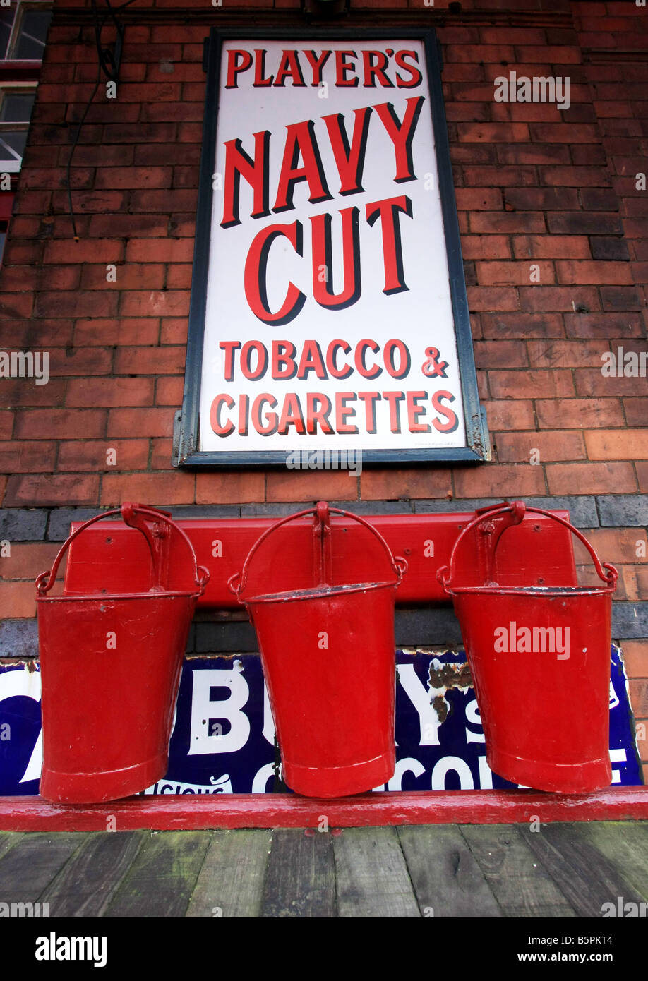 Three (3) old fashioned red fire buckets beneath an old enamel sign advertising Players Navy Cut cigarettes Stock Photo