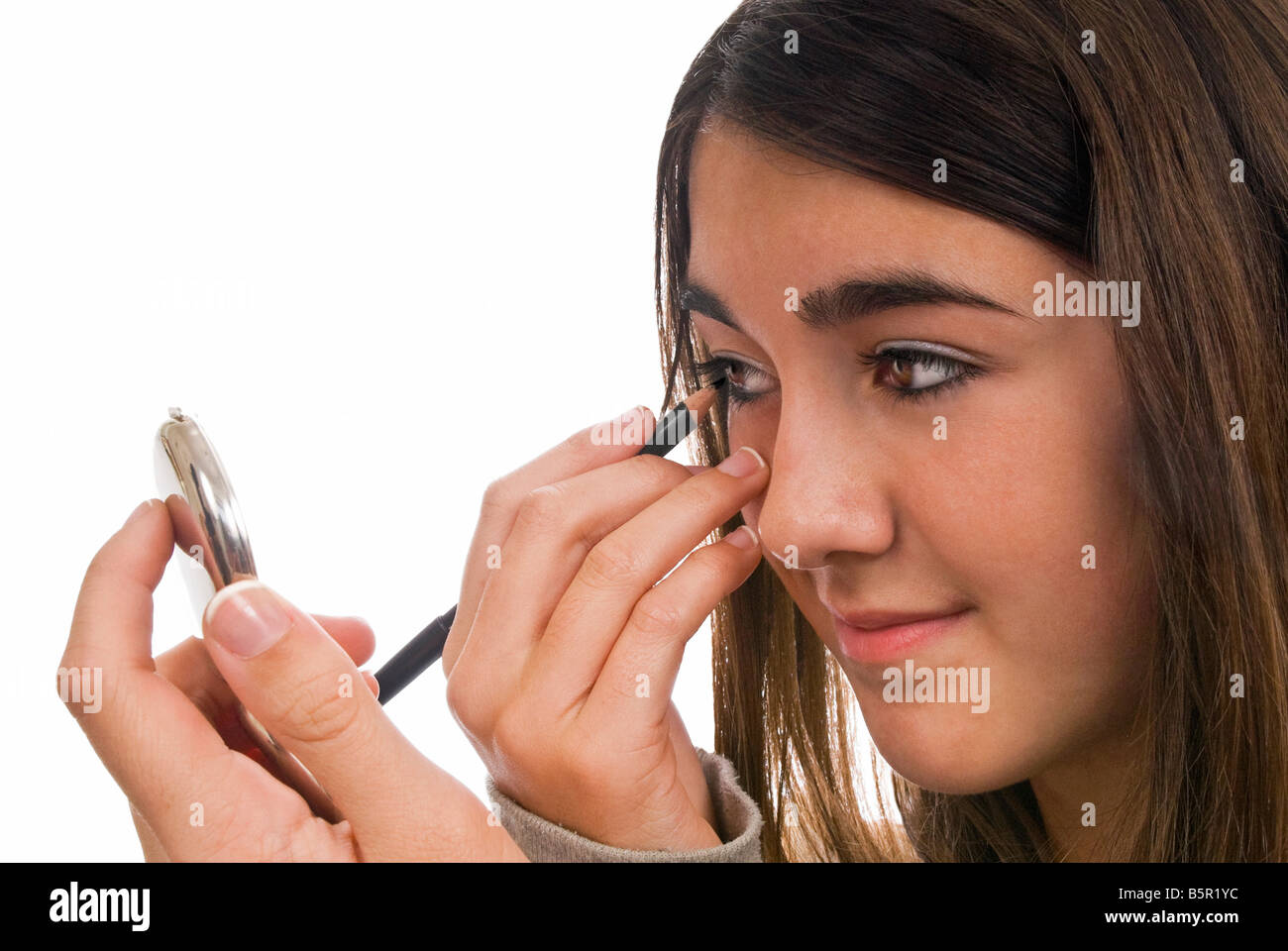 Horizontal portrait of a young girl applying black kohl eyeliner around her eyes in a compact mirror against a white background Stock Photo