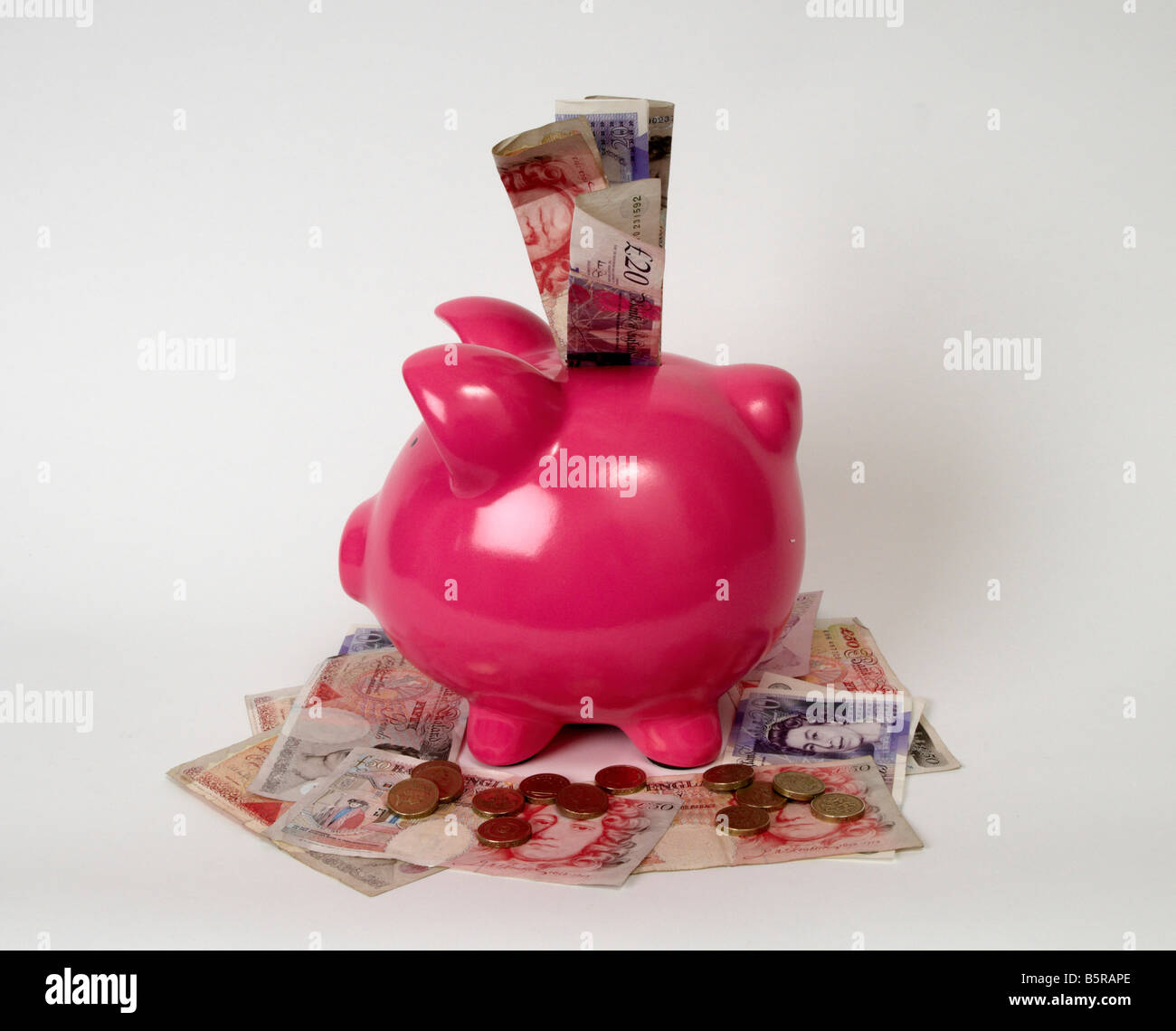 profile of a pink piggy bank sitting on a pile of british bank notes and coins with bank notes sticking out the top of it. Stock Photo