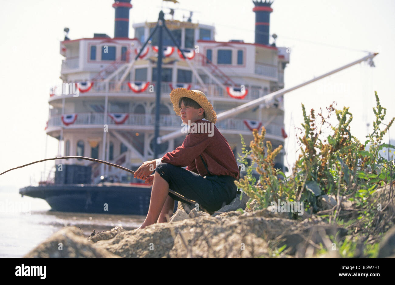 A Huck Finn Huckleberry Finn look alike fishes for catfish on the Mississippi River with the Mississippi Queen steamboat, Hannibal, Missouri. Stock Photo