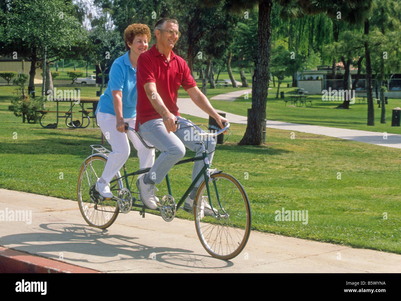 Senior couple ride tandem bicycle built for two in park. Stock Photo