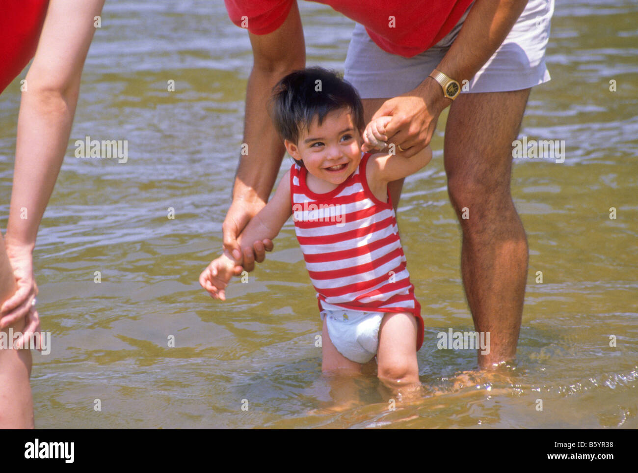 Hispanic toddler laughs as his parents help him wade in lake waters. Stock Photo