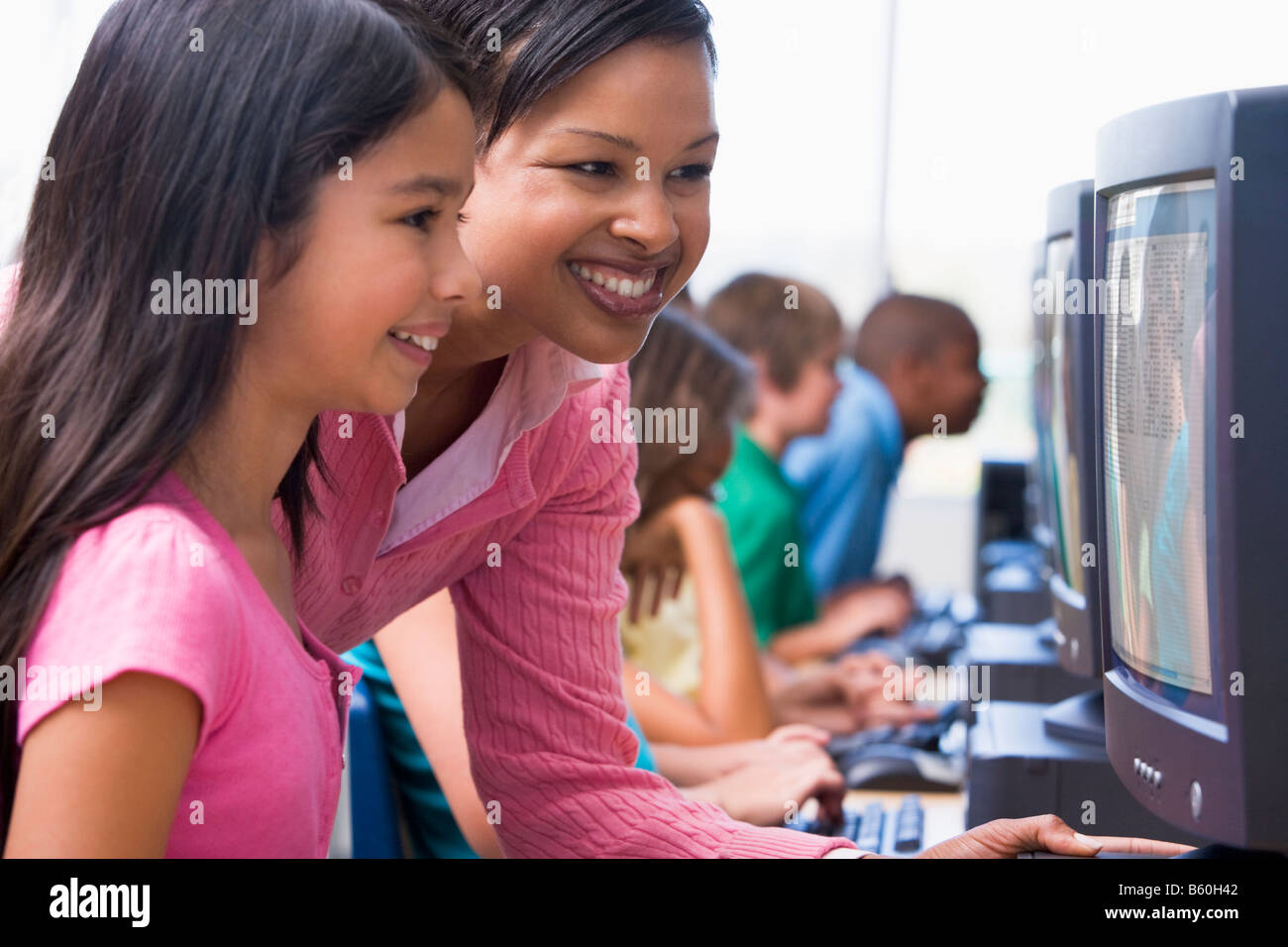 Teacher helping student at computer terminal with students in background (depth of field/high key) Stock Photo