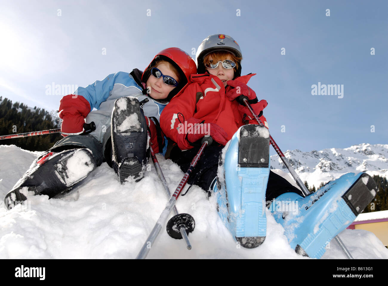 Two children wearing ski boots, helmets and sunglasses sitting on a