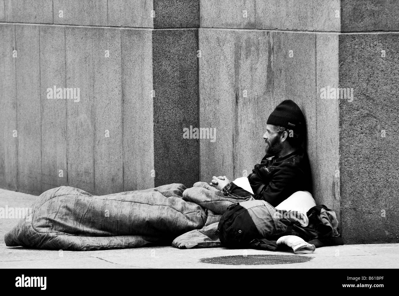 A black and white photo of a homeless man, a bum, a beggar, sitting on the street corner of a street in a major city. Stock Photo