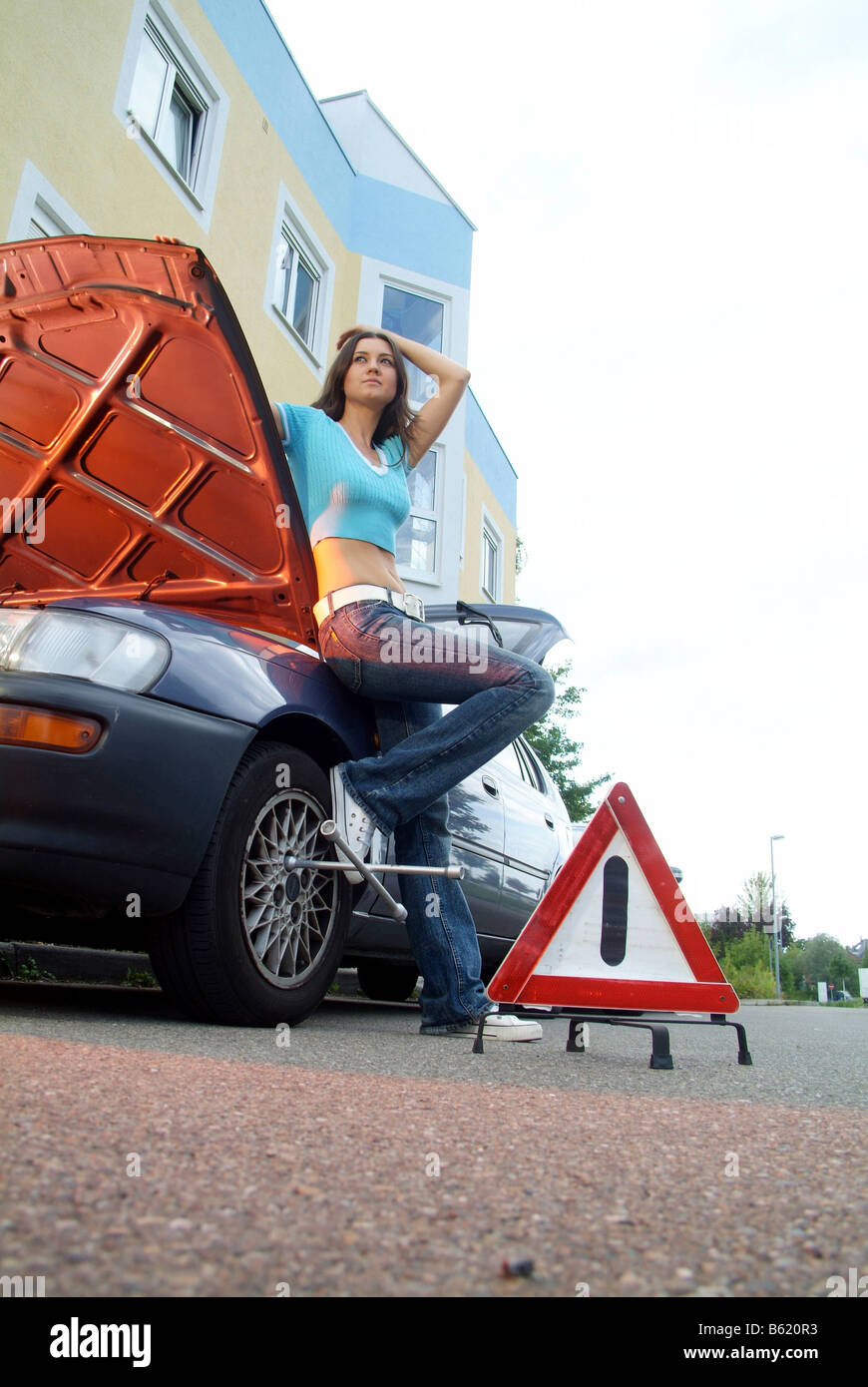 Young woman next to a warning triangle, leaning against her broken down car and waiting for help Stock Photo
