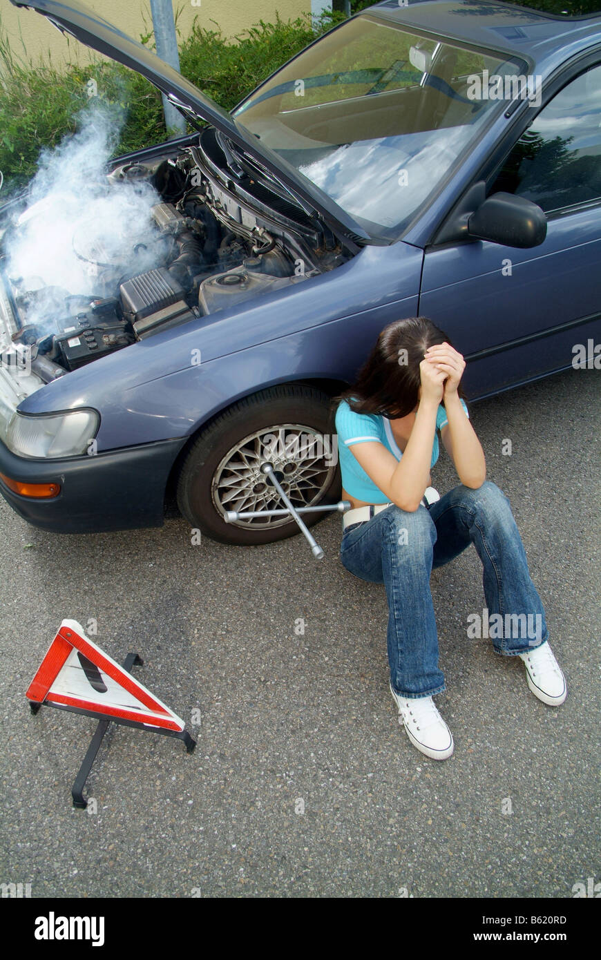 Frustrated young woman with a warning triangle sitting next to her broken down and smoking car and waiting for help Stock Photo