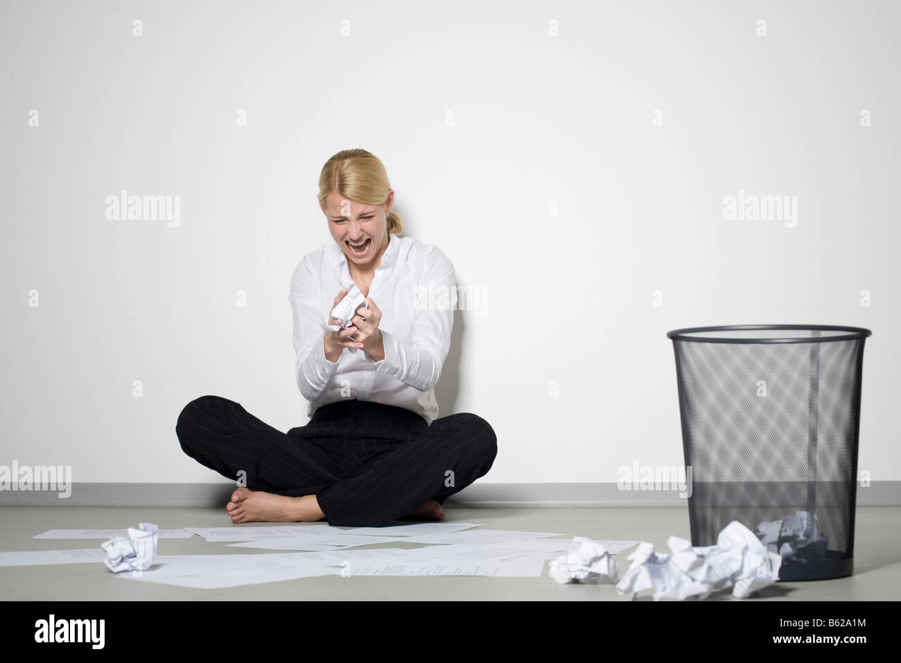 Young blonde woman sitting by the wall in despair amongst crumpled paper Stock Photo