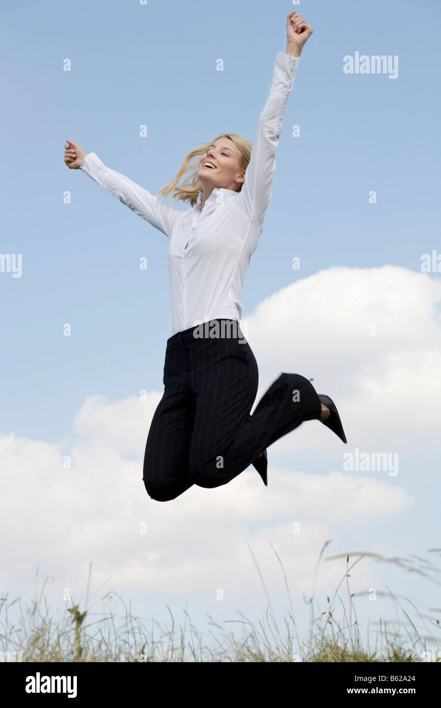 Young blonde woman wearing business clothing jumping in front of a blue sky, outstretched arms Stock Photo