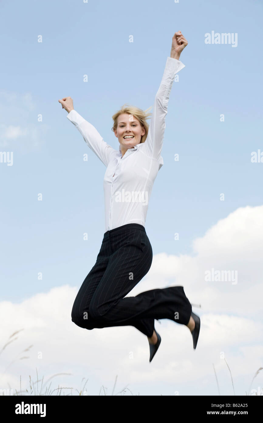 Young blonde woman wearing business clothing jumping in front of a blue sky, outstretched arms Stock Photo