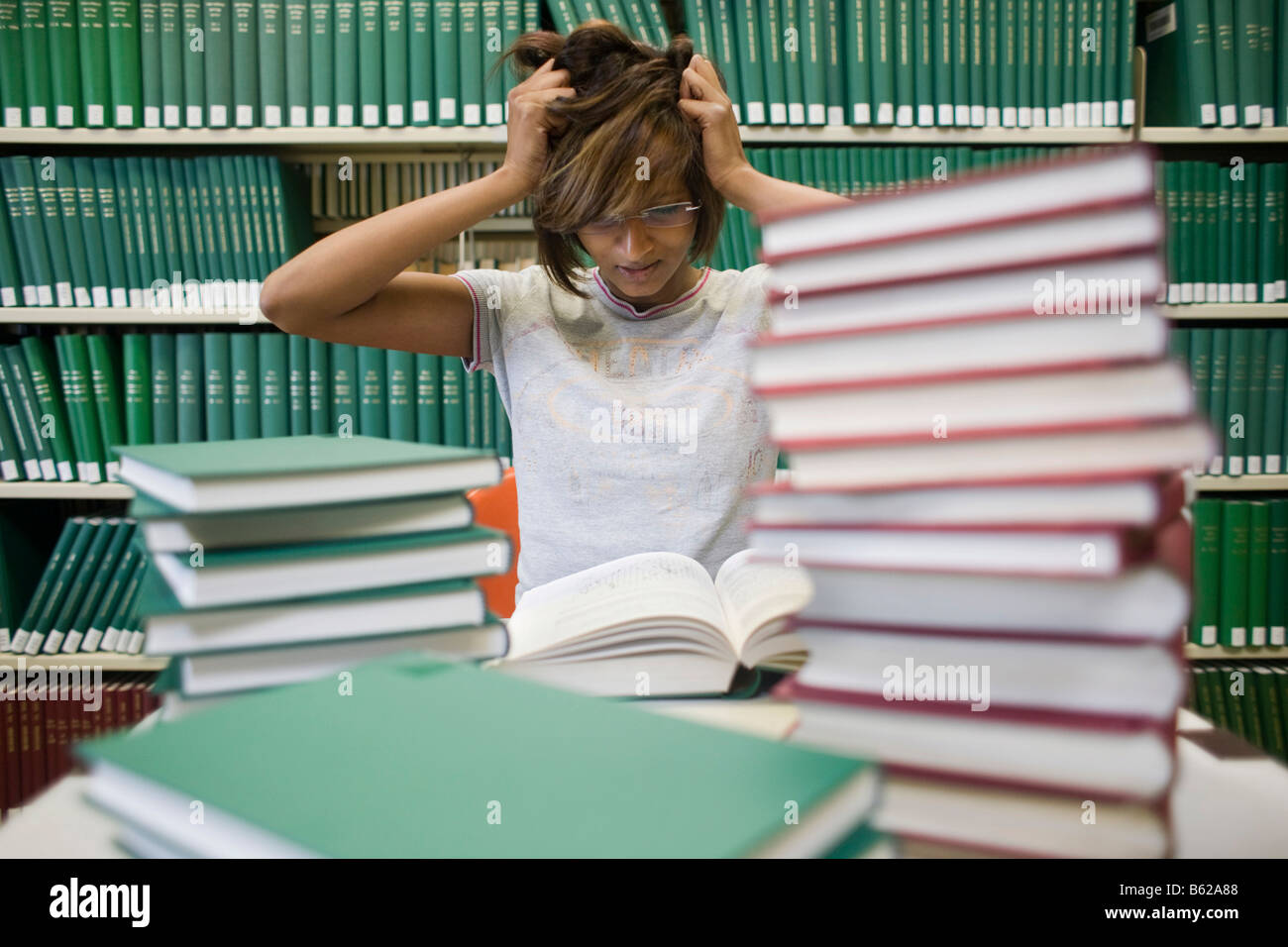 Young dark-haired female student sits despairingly with many books at a table in front of a bookshelf in a library Stock Photo