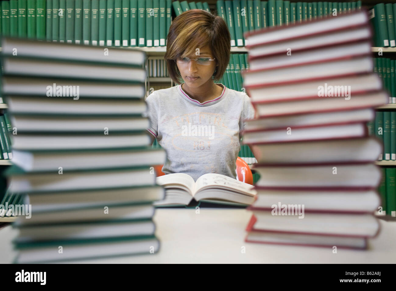 Young dark-haired female student sitting with many books at a table in front of a bookshelf in a library Stock Photo