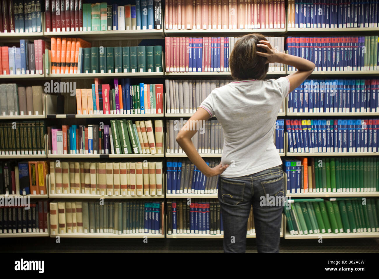 Young dark-haired female student standing helplessly in front of a bookshelf in a library Stock Photo