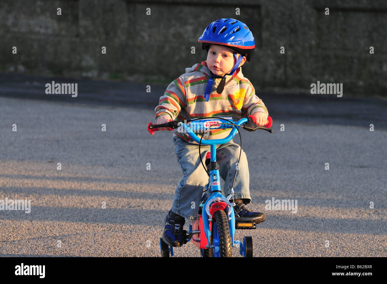 Boy [3 years] infant riding bicycle bike stabilizers [hard work ]effort pushing peddles hard hat helmet Stock Photo