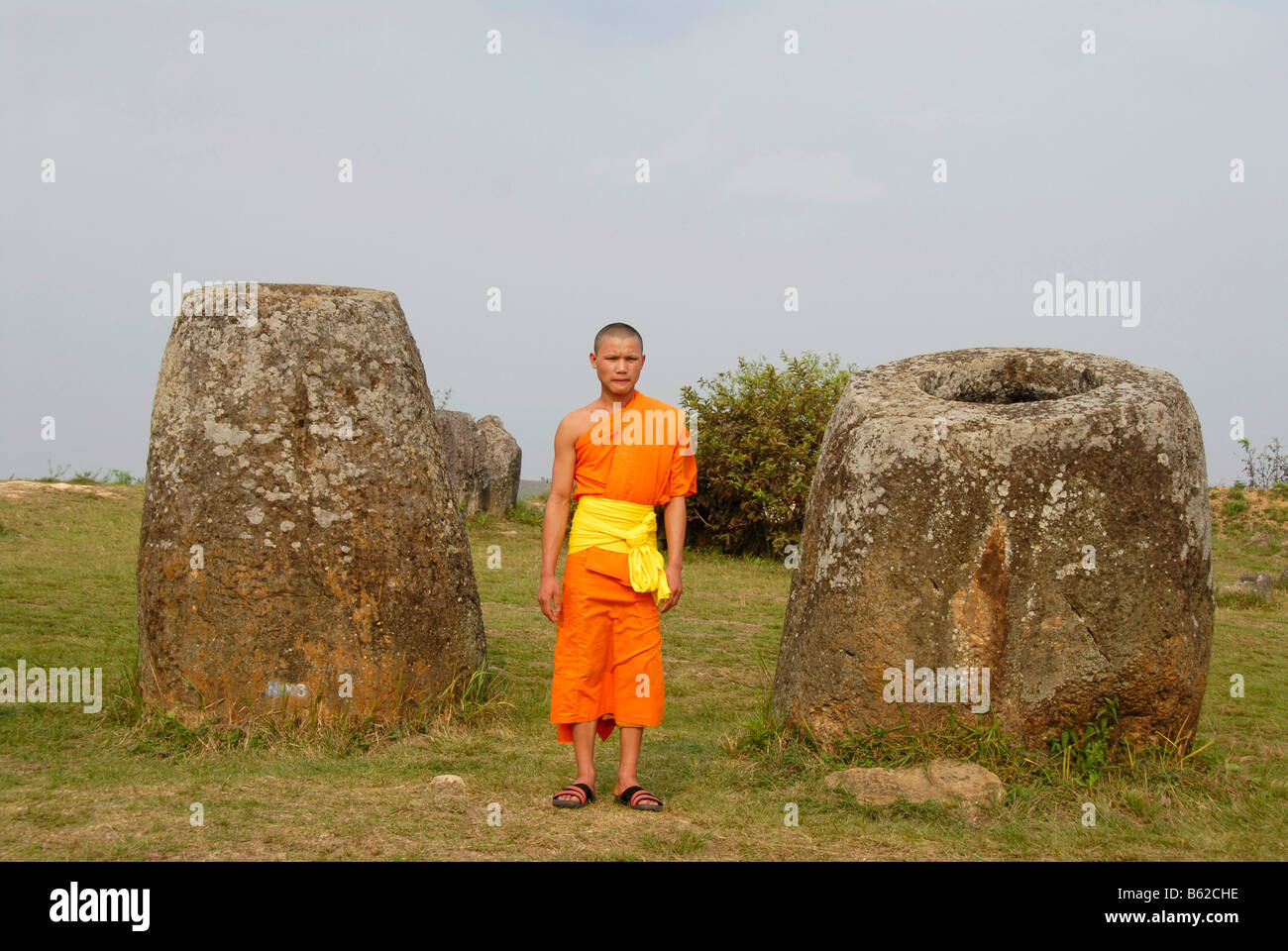 Young monk wearing an orange cowl standing between enormous jars, monoliths made of stone, Plain of Jars, Xieng Khuang Province Stock Photo