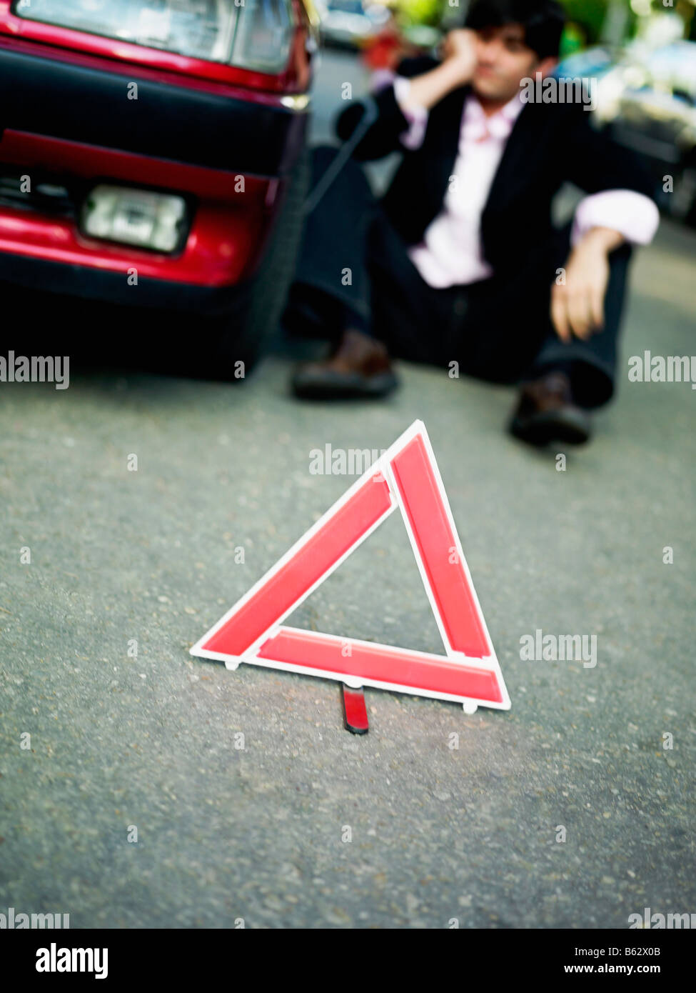Mid adult man sitting by a broken-down car behind a warning triangle on the road Stock Photo