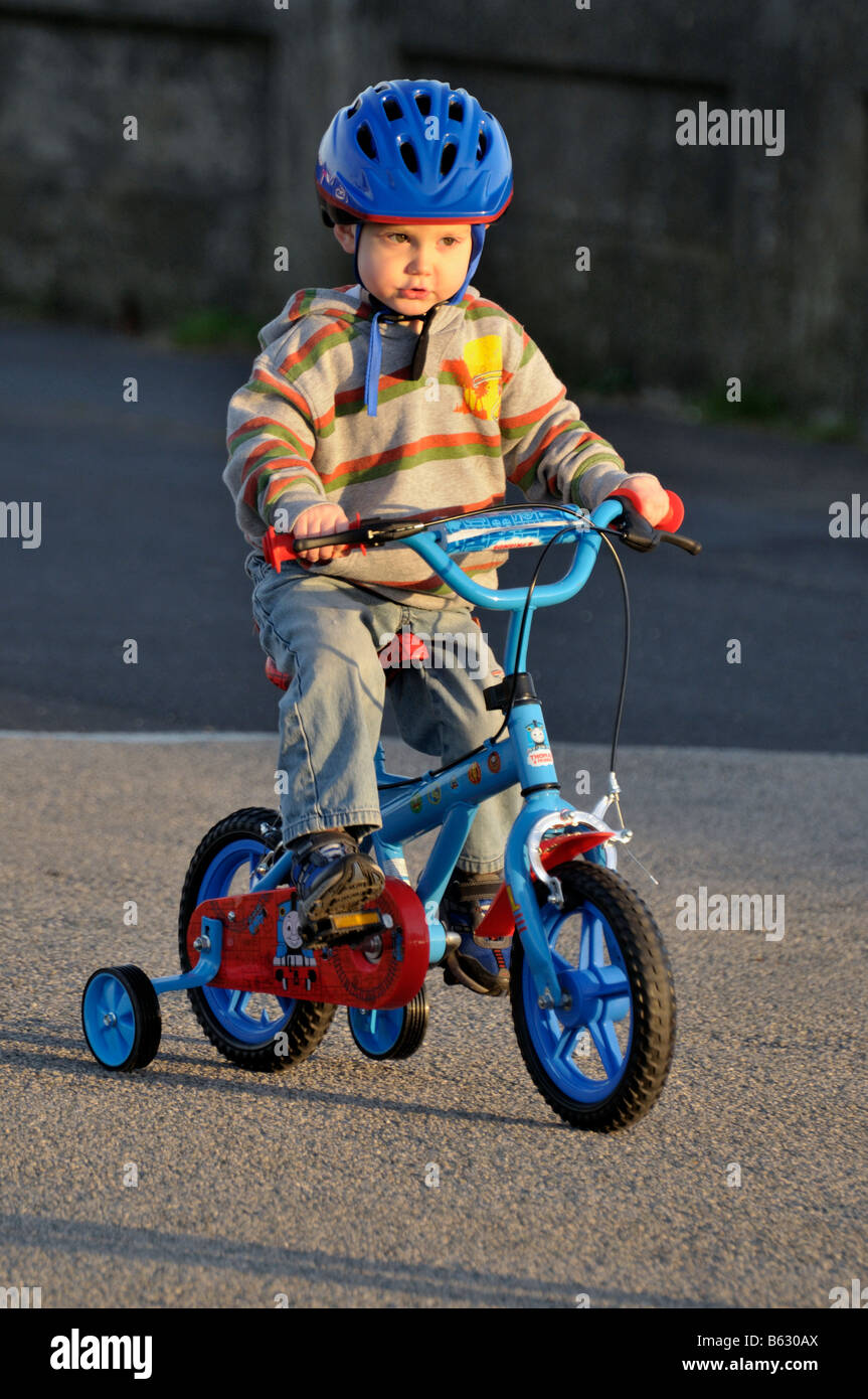 Boy [3 years] infant riding bicycle stabilizers bike [ hard work] effort pushing peddles hard hat helmet outdoors play Stock Photo